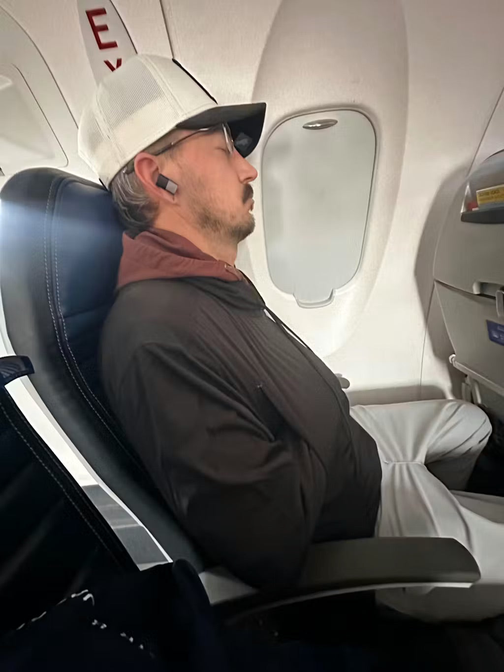 Man sleeping upright in an airplane seat wearing the STR-8 Jacket with both arms resting inside the built-in chest support pocket